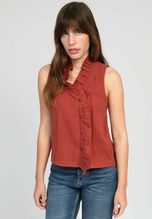 Femme aux longs cheveux bruns portant un chemisier sans manches rouge rouille à volants sur la patte de boutonnage, associé à un jean bleu, sur un fond clair uni.