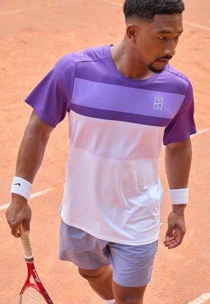 Lila und weiße Sportshirt mit kurzen Ärmeln, das horizontale Streifen aufweist. Getragen mit grauen Shorts und hält einen roten Tennisschläger.