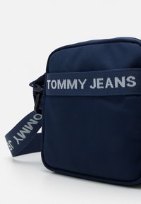 Bolso bandolera rectangular azul marino hecho de tela texturizada, con una correa blanca con el logo "TOMMY JEANS" y cintas ajustables para llevar.