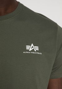 Camiseta de algodón verde oliva con cuello redondo y un gráfico de logo en blanco que incluye elementos geométricos y texto.