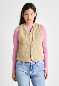 Kaki bomullsvest med to frontlommer, rundet hem og trykknapper, brukt over en rosa ribbestrikket genser og lyseblå jeans.