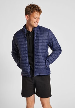 Marineblå pufferjakke med quiltet design, høj krave, lynlåslukning og sidelommer. Båret over en sort polo og shorts. Glat tekstur.