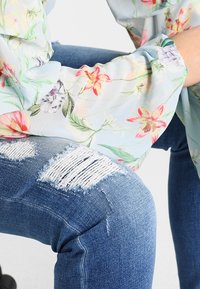 Henkilö, joka on pukeutunut revittyihin sinisiin farkkuihin ja vaaleansiniseen kukalliseen pitkähiharaiseen puseroon, istuu yksi polvi koukistettuna.