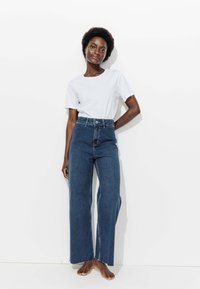 Jean en denim bleu foncé taille haute, avec une coupe décontractée et des jambes larges, associé à un t-shirt blanc à col rond simple. Détails minimaux.