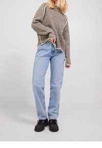 Personne portant un pull à rayures marron et blanches, un jean bleu clair et des chaussures noires, debout sur un fond gris clair uni.