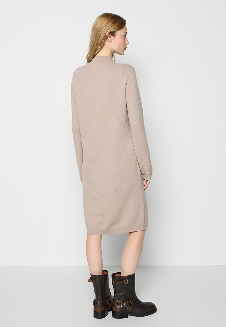 Beige Strickkleid mit langen Ärmeln, knielang, mit hohem Kragen. Kombiniert mit dunkelbraunen Stiefeletten mit Schnallenakzenten.