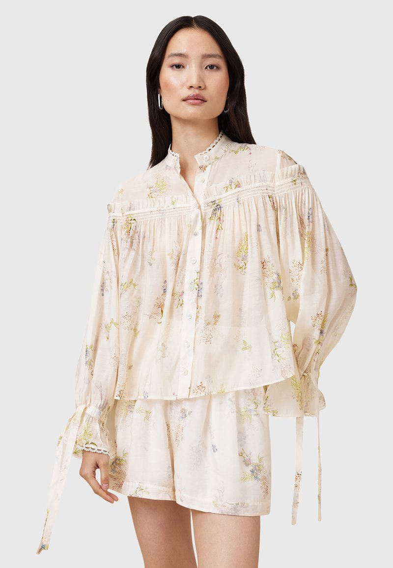Blusa floreale leggera in crema con maniche ampie e chiusura a bottoni, con rifiniture in pizzo e dettagli arricciati. Abbinata a pantaloni corti coordinati.