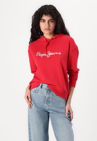Sarkans sporta krekls ar baltu "Pepe Jeans London" logo, ar regulējamu kapuci un ribinātām piedurknēm, apvienots ar gaiši ziliem džinsiem.