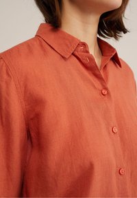 Leinenhemd in Rostfarbe mit Kragen, ausgestattet mit fünf Knopfverschlüssen, subtiler Textur und kurzen Ärmeln.