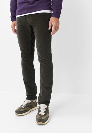 Mand iført mørkegrønne kordbuksersbukser, grønne og beige sneakers med hvide snører og en lilla sweater.