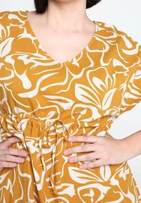 Żółta sukienka z białym abstrakcyjnym wzorem kwiatowym, dekoltem w serek i krótkimi rękawami. Posiada sznurki w talii umożliwiające regulację dopasowania.