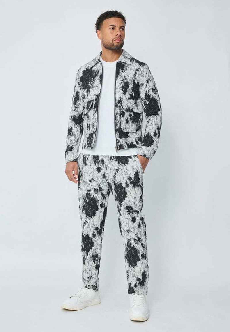 Homme portant un ensemble coordonné de veste et pantalon à imprimé abstrait noir et blanc, T-shirt blanc et baskets blanches, se tenant avec une main dans la poche.