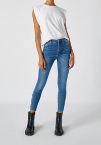 Jeans skinny azules de cintura alta y acabado desgastado, combinados con una camisa sin mangas blanca y botines negros con suelas gruesas.