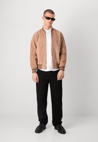 Suede Bomberjacke in hellbraun mit gerippten Bündchen und grünen sowie roten Akzenten, getragen über einem weißen Hemd mit Knöpfen und schwarzen Hosen.