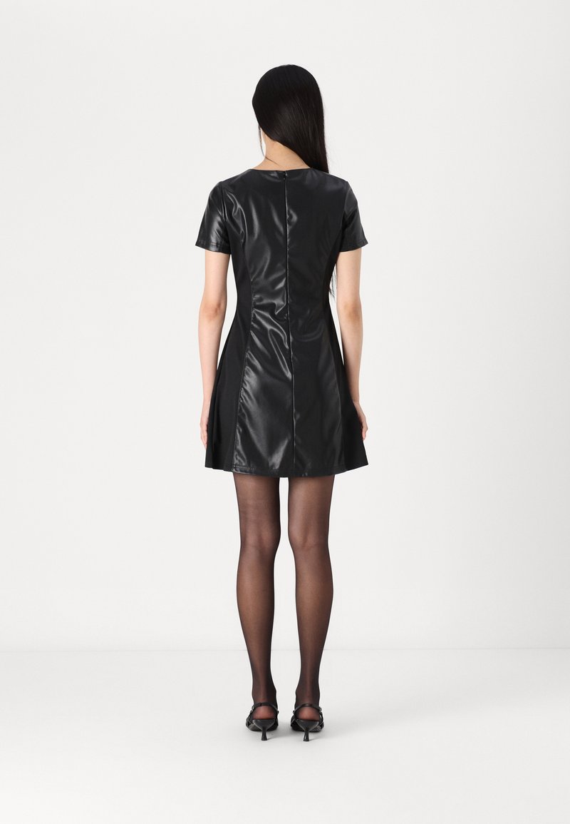 Schwarzes Kunstlederkleid mit kurzen Ärmeln, tailliertem Oberteil, ausgestellt Rock und Reißverschluss auf der Rückseite. Kombiniert mit transparenten schwarzen Strumpfhosen und Schuhen mit Absatz.