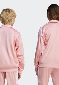Rosa sateng treningsdresser med ribbestrikkede mansjetter og hem. To modeller vises bakfra, iført matchende antrekk med hvite sidestreker.
