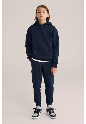 Niño con cabello de longitud media, de pie, vistiendo una sudadera con capucha azul marino, pantalones cargo jogger a juego y zapatillas deportivas blancas y negras.