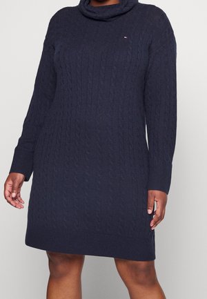 Dark blue cable-knit long-sleeve sweater dress with turtleneck, ribbed cuffs, and small red, white, and blue embroidered logo on chest.