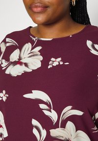 Top bordeaux con un motivo floreale; presenta fiori bianchi sovradimensionati con accenti rossi. Tessuto liscio e leggero con scollatura a girocollo.