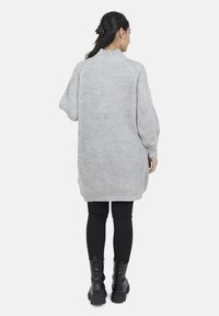 Grå strikket sweater med høj krave og løs pasform, præget af ribbet tekstur. Båret over sorte leggings og kampstøvler.