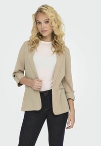 Beige blazer van een gestructureerde stof, met een enkele knoop op opgerolde mouwen, voorzakken en een open voorkant, gecombineerd met een licht shirt en donkere jeans.
