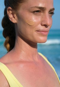 Modella con capelli castano chiaro, indossa un costume da bagno giallo, ha un'abbronzatura e lucentezza sulla pelle, con una base beige applicata sulla guancia. Oceano sullo sfondo.