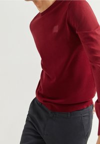 Czerwony sweter z dzianiny, okrągłym dekoltem i długimi rękawami. Posiada małą naszywkę z logo na piersi oraz ściągacze przy rękawach i u dołu.