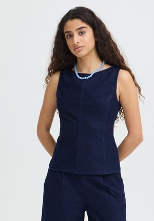 Žena s dlhými kučeravými vlasmi, ktorá nosí tielko námorníckej modrej farby, zodpovedajúce nohavice a svetlomodrý náhrdelník z korálikov, stojí pred jednoduchým pozadím.