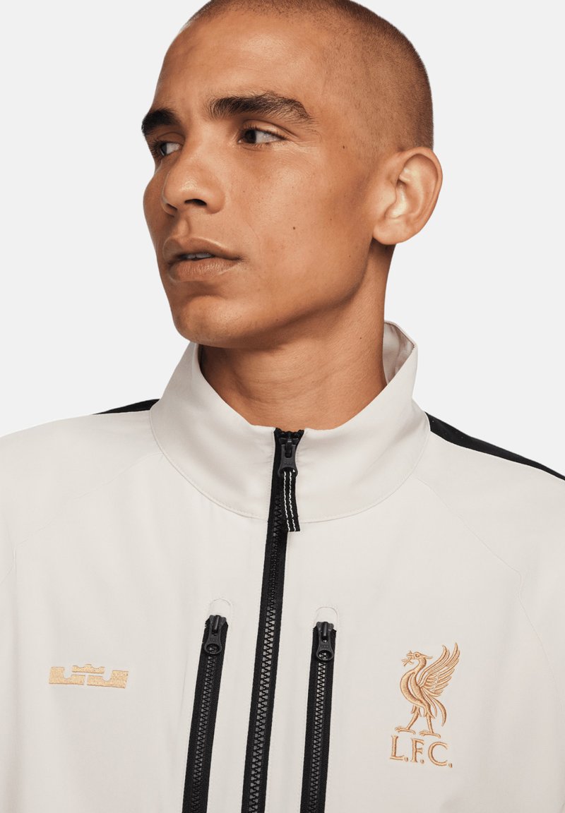 Chaqueta deportiva blanca con cuello alto, acentos negros, bolsillos con cremallera y logotipos en relieve de L.F.C. y LeBron James en dorado. Textura suave.