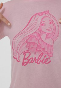 Růžové bavlněné tričko s karikaturou ženské postavy s dlouhými vlasy a náušnicemi ve tvaru srdce, s nápisem "Barbie" pod obrázkem.