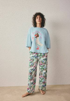 Žena s kučeravými vlasmi, oblečená v svetlomodrej voľnej košeli s potlačou kačice a farebných tropických nohaviciach, stojaca naboso pri hladkej stene.