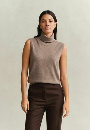 Vrouw met lang donker haar, die een mouwloze beige coltrui en donkerbruine broek draagt, staand tegen een effen lichte achtergrond.