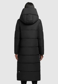 Kvinde iført en lang sort dunjakke med en stor hætte, set bagfra mod en ensfarvet grå baggrund.
