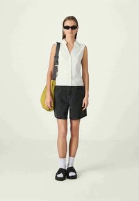 Witte mouwloze blouse, zwarte denim shorts, gele tas met ribbelstructuur, zwarte sandalen en witte sokken; minimale accessoires en strakke lijnen.