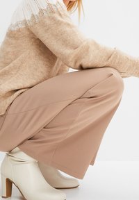 Beiger Strickpullover mit weißem Spitzenmuster an den Schultern, kombiniert mit hellbraunen weit geschnittenen Hosen und weißen Blockabsatz-Stiefeletten.