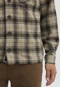 Chemise en flanelle à carreaux beiges avec des checks marron et bleu marine, présentant un devant à bouton et des poignets. La chemise a une texture douce, présentée avec un pantalon marron.