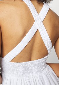 Bílé šaty s křížovými ramínky. Mají strukturovaný, smockovaný elastický pás v pase a hladkou látku na zadní straně.