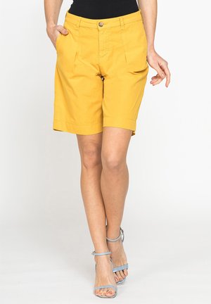 Persona che indossa pantaloncini gialli fino al ginocchio, top nero e sandali con tacco azzurri, in piedi con una mano in tasca.
