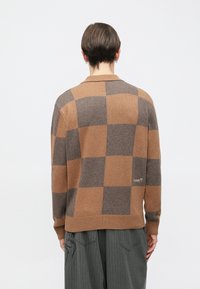 Smeđi i bež pulover na kockice s dugim rukavima, jednobojnim ovratnikom, rebrastim rubom i vezanim logotipom na leđima.