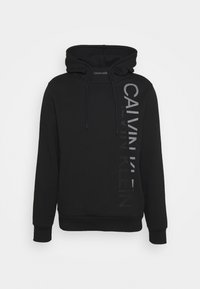 Černá mikina s kapucí vyrobená z měkkého materiálu, s šňůrkou na stažení. Na přední straně se nachází vertikální, reflexní logo Calvin Klein.