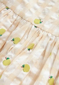 Robe en coton beige clair avec un motif à carreaux, ornée de pommes jaunes brodées avec des feuilles vertes, et des plis froncés le long de la taille.