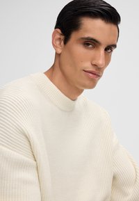 Mann mit dunklem, zurückgegeltem Haar, der einen weißen gerippten Strickpullover trägt und in die Kamera schaut, vor einem schlichten hellgrauen Hintergrund.