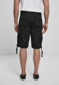 Homme portant un short cargo noir et des baskets noires, se tenant de dos sur un fond gris uni.