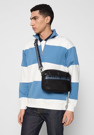 Blau-weiß gestreifter Pullover mit Reißverschluss am Kragen, kombiniert mit einer schwarzen Umhängetasche mit blauem Logoband. Dunkle Denim-Jeans.