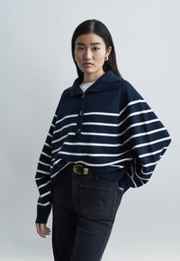 Jeune femme aux longs cheveux noirs portant un pull à rayures bleu marine, un jean noir, une boucle de ceinture dorée et des boucles d'oreilles créoles dorées, se tenant devant un fond gris.