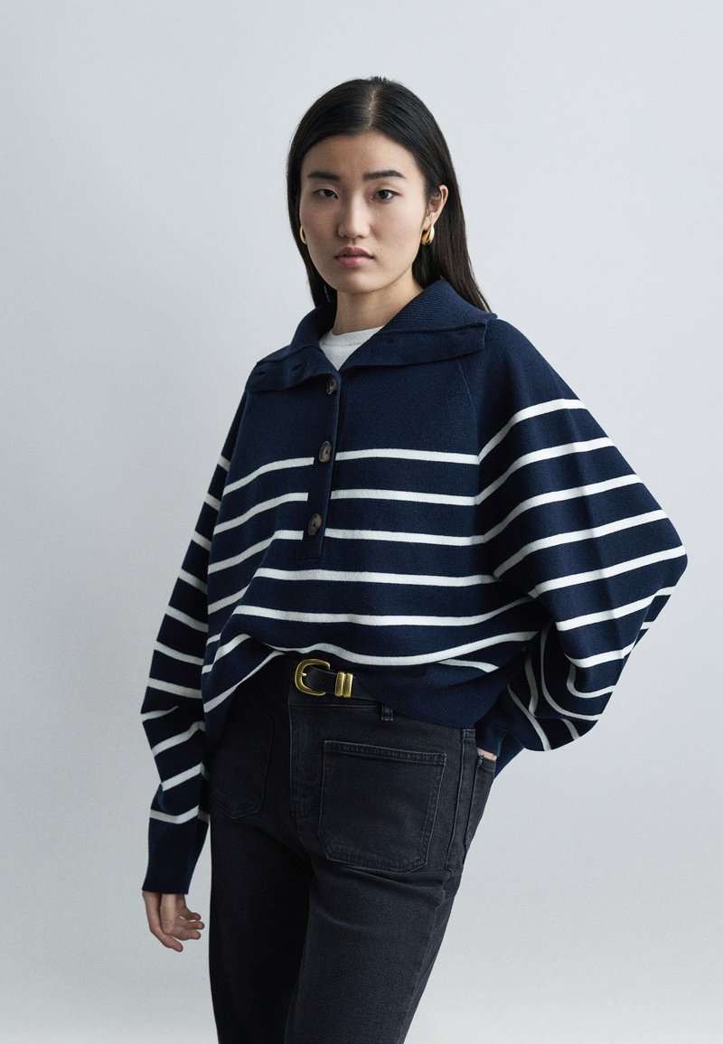 Jeune femme aux longs cheveux noirs portant un pull à rayures bleu marine, un jean noir, une boucle de ceinture dorée et des boucles d'oreilles créoles dorées, se tenant devant un fond gris.