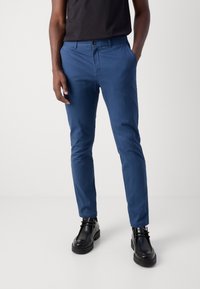 Chinos de mezcla de algodón azul con un ajuste slim, que cuentan con cierre de botón, bolsillos laterales y una textura suave, combinados con zapatos de cuero negros.