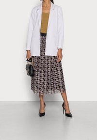 Blazer blanc sur un top décontracté brun, associé à une jupe plissée à motif géométrique en violets et noirs, et des talons hauts noirs.