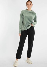 Groene fluwelen top met lange mouwen en een ronde hals, gecombineerd met zwarte rechttoe-rechtaan spijkerbroek en beige sneakers. Soepele textuur, casual pasvorm.