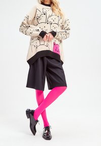 Femme portant un pull beige orné de visages de chats noirs et d'un visage de chat rose, un short large noir, des collants rose vif et des chaussures noires brillantes à lacets.